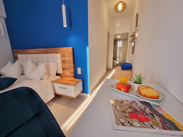 A modern bedroom with a blue accent wall, a wooden headboard, a bedside table with a lamp, and a breakfast tray with fruit and bread. - Hotel Nefeli (Volos)