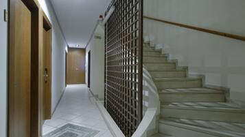 Um corredor com uma porta de madeira à esquerda, uma escada com corrimão de metal à direita e piso ladrilhado.