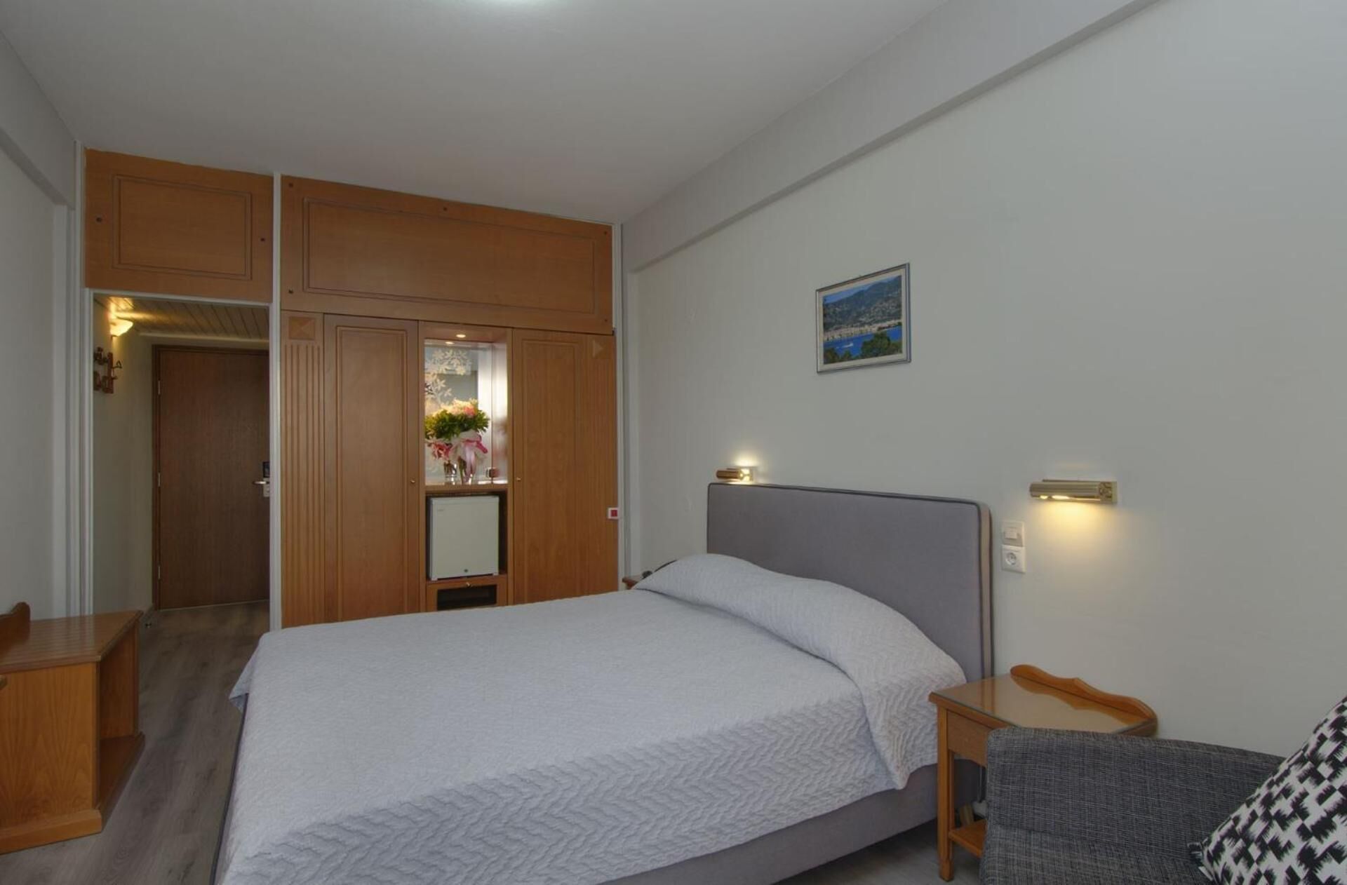 Een slaapkamer met een bed, een houten garderobe, een bureau en een stoel. Er staat een klein tafeltje met een lamp en er hangt een prent aan de muur.