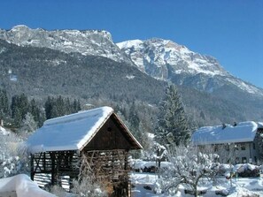 Une cabane en bois, dont le toit est recouvert de neige, entourée d’arbres chargés de neige et avec des montagnes en arrière-plan.