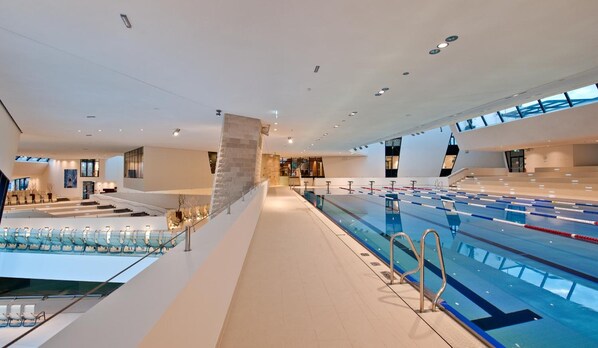 Une piscine intérieure moderne dotée de plusieurs couloirs, d’un espace détente avec des chaises et d’un escalier menant à un niveau supérieur.