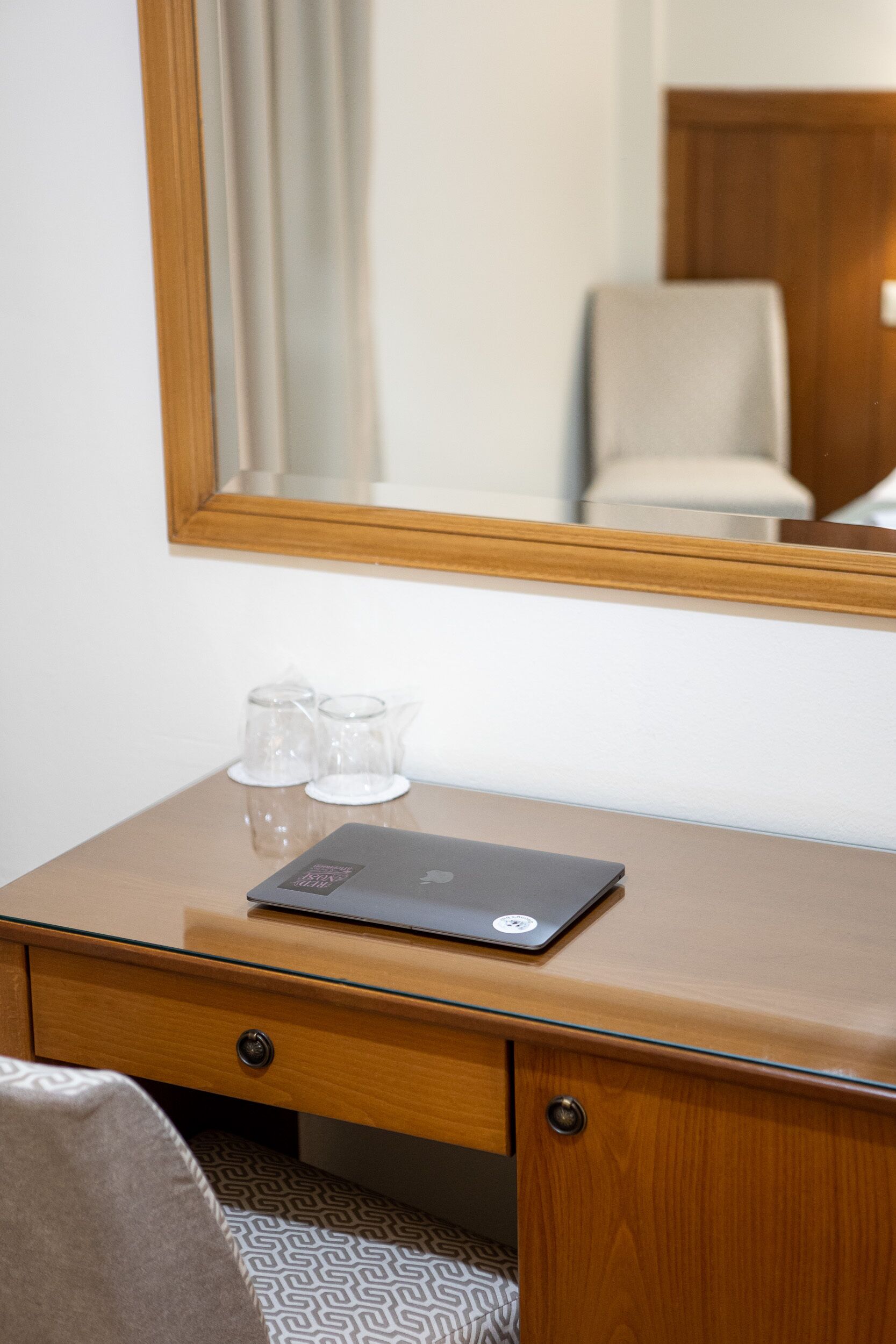 A wooden desk with a laptop, two clear glasses, and a mirror reflecting a chair and curtains.