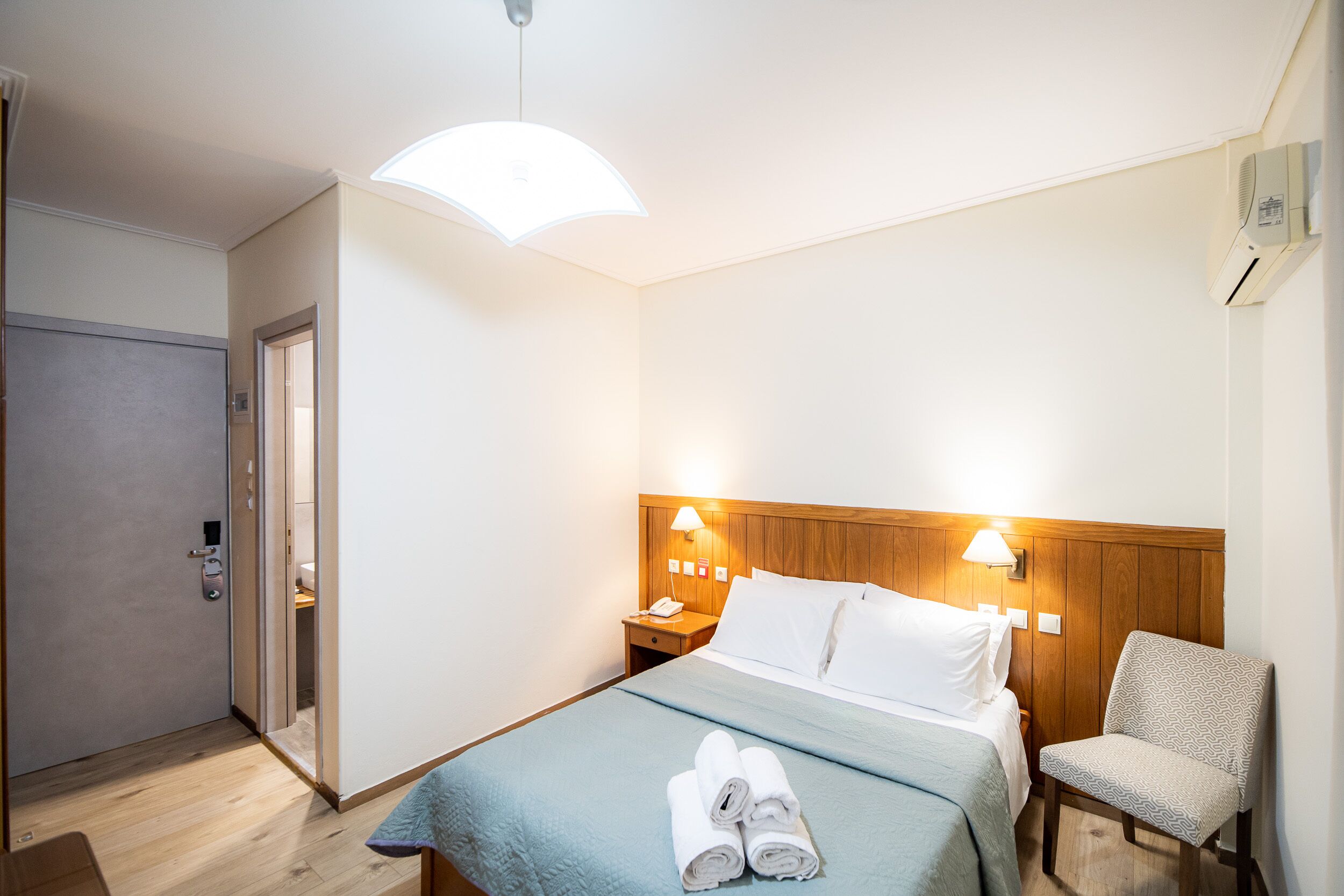 A hotel room with a bed, a chair, a nightstand, and a door.