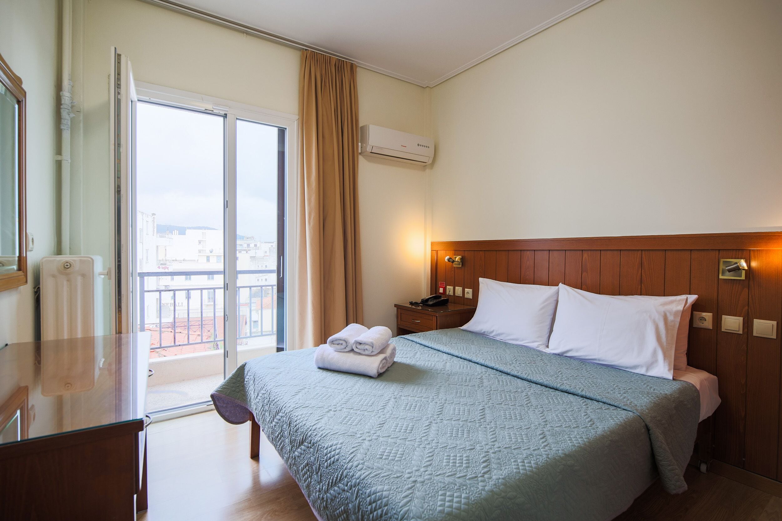 A hotel room with a bed, a desk, a window with curtains, and an air conditioning unit.