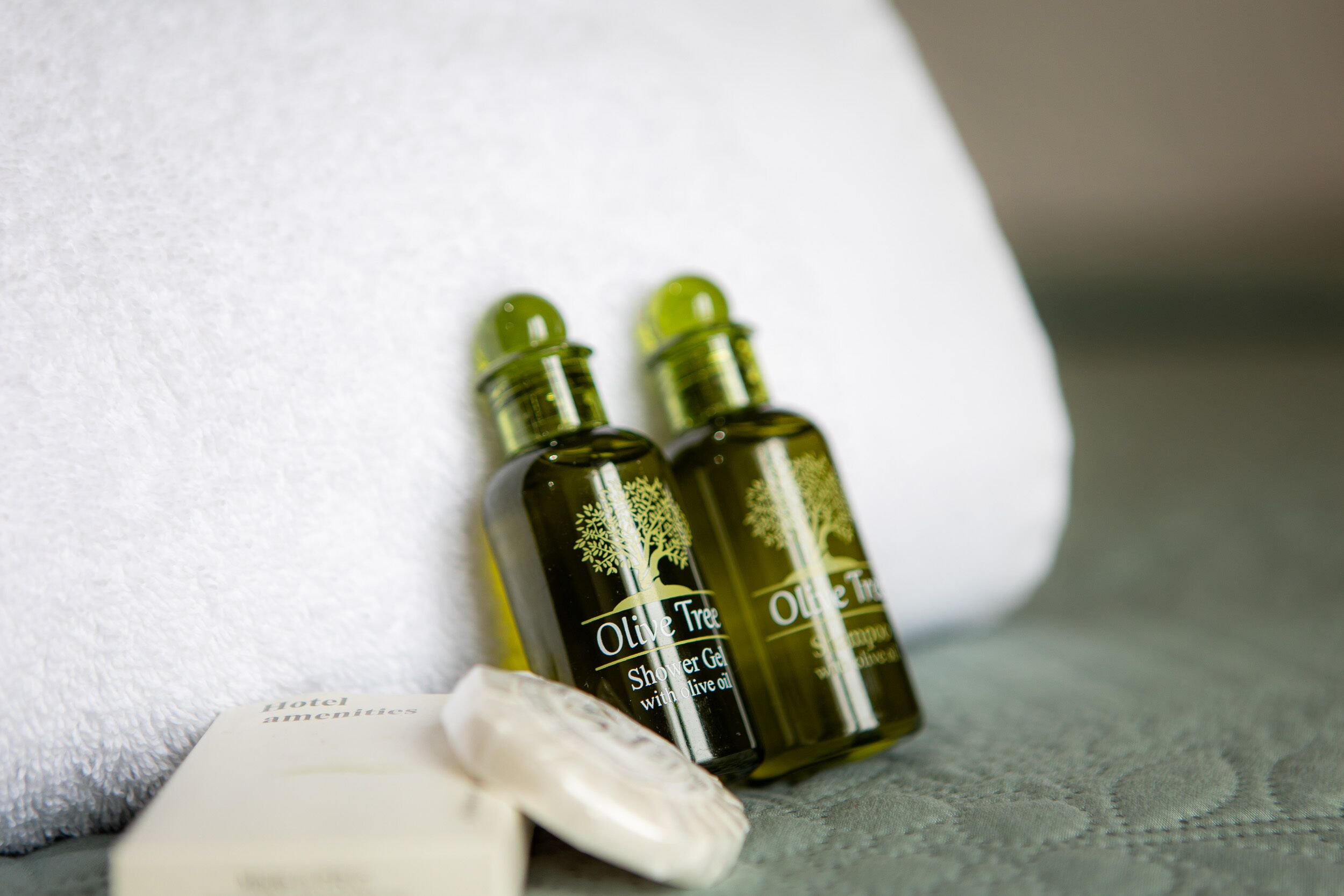 Two bottles of Olive Tree Shower Gel with olive oil, a bar of soap, and a white towel on a textured surface.
