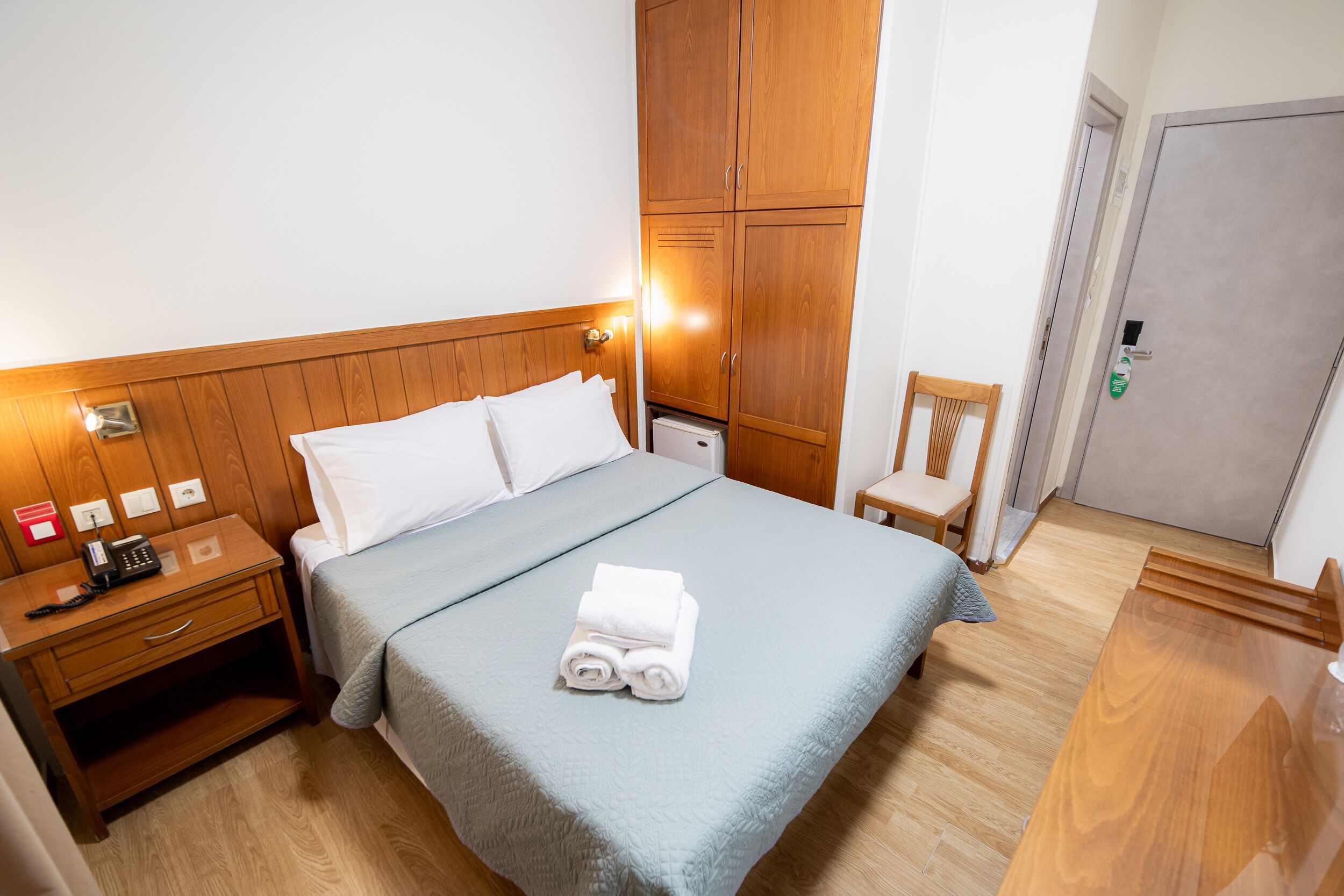 Een netjes opgemaakt bed met een blauwe sprei, een houten nachtkastje met een telefoon en een lamp, en een houten garderobe.