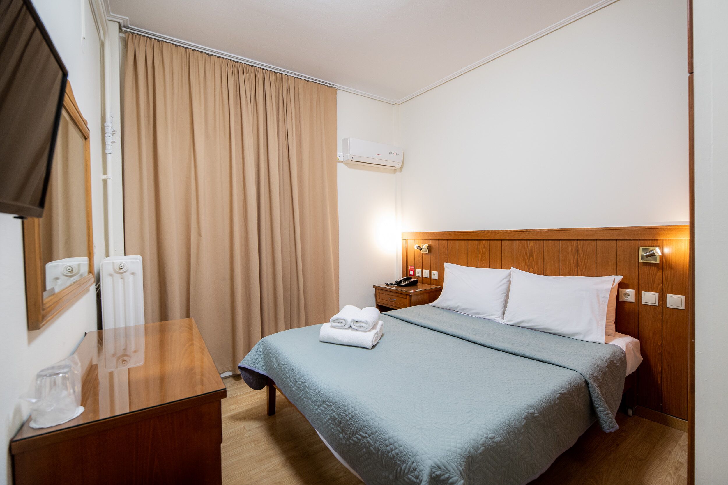 A hotel room with a bed, a wooden headboard, a nightstand, a mirror, and a window with curtains.