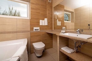 A modern bathroom with a bathtub, toilet, and sink, featuring a large mirror and a window with a view of trees.