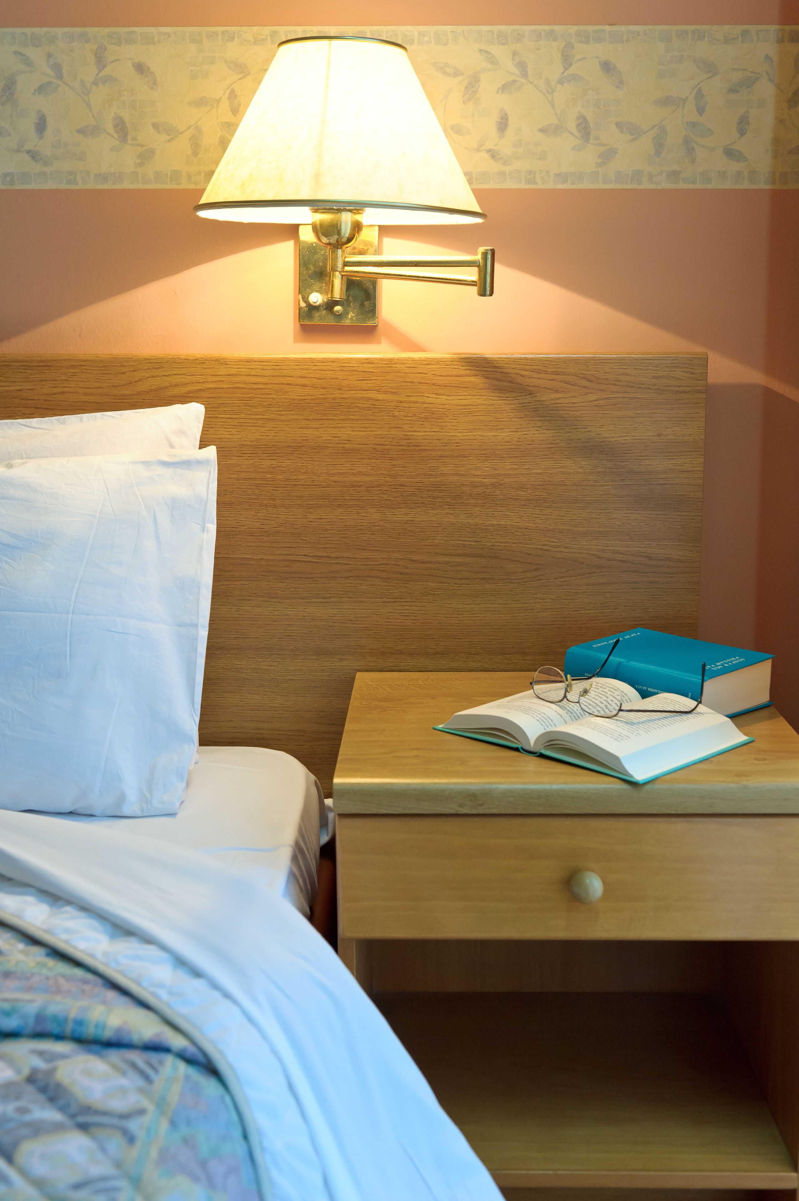 A neatly made bed with a pillow, a bedside lamp, a book, and glasses on a nightstand.