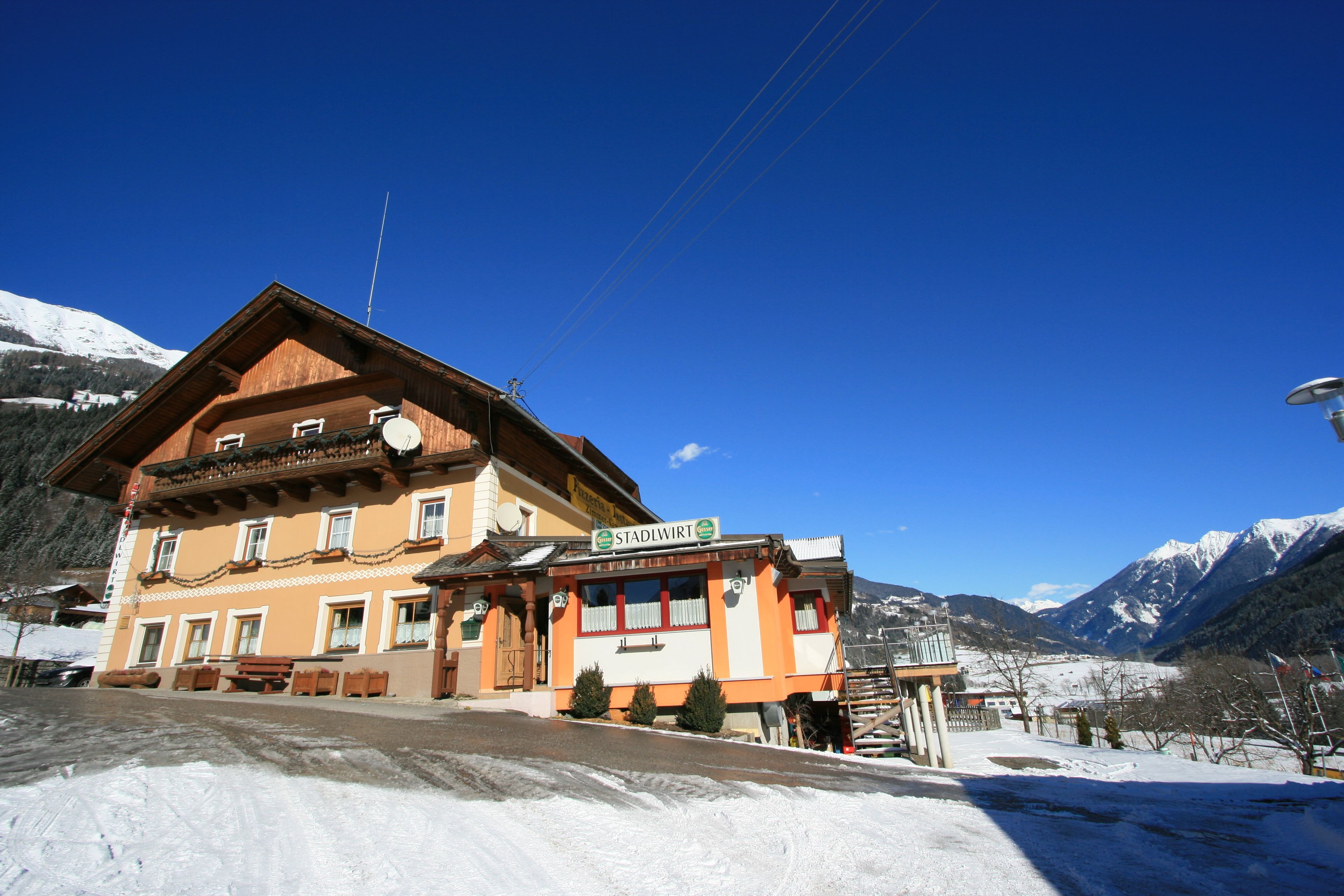 Ein traditionelles Chalet mit einem Schild, auf dem "Stadlwirt" steht. Es hat eine Holzfassade, mehrere Fenster und einen Balkon. Die Umgebung ist verschneit mit Bergen im Hintergrund.