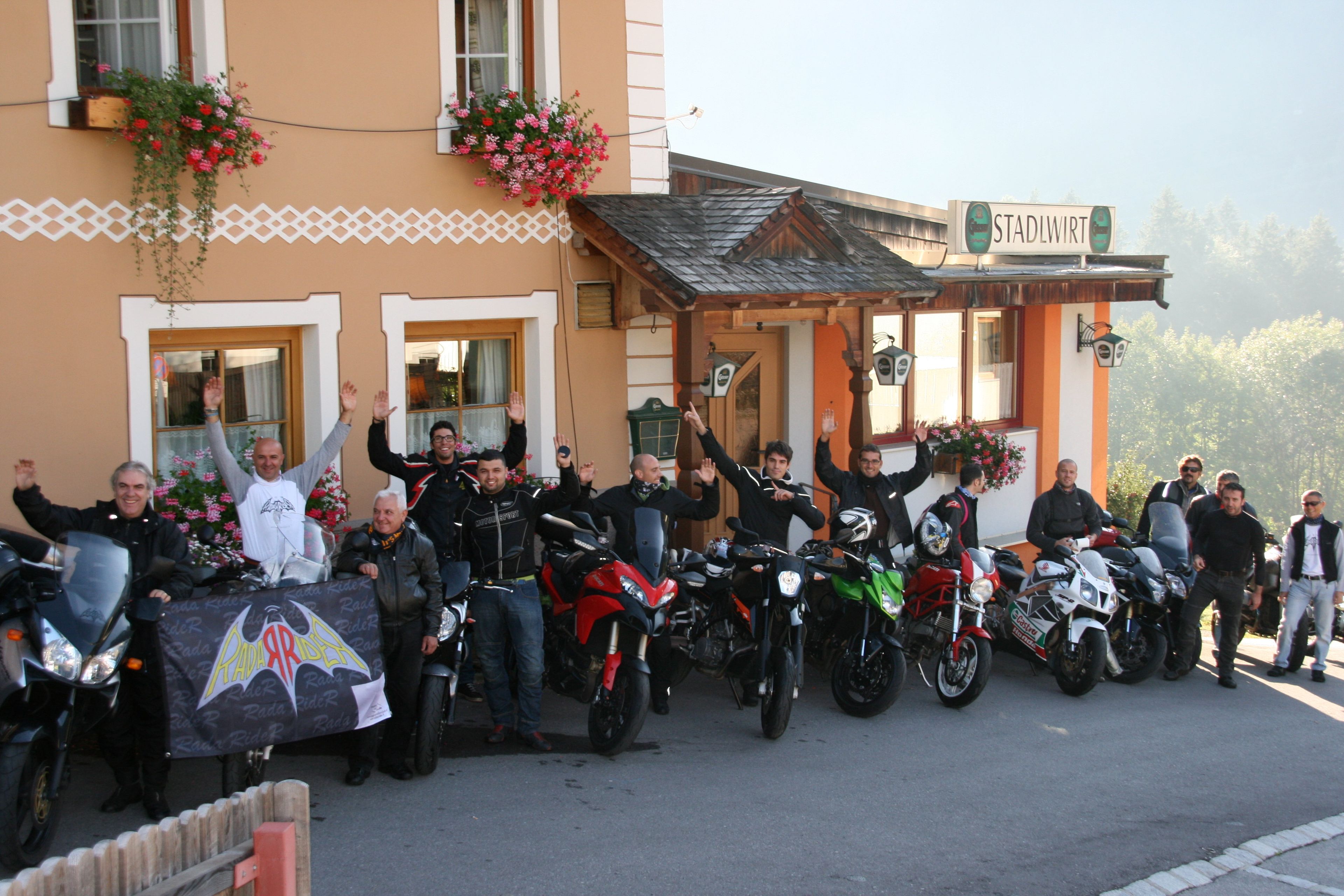 Eine Gruppe von Personen mit Motorrädern vor einem Gebäude, an dem ein Schild mit der Aufschrift 'STADLWIRT' hängt.