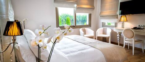 A hotel room with a bed, a desk, two chairs, a TV, and a window with a view of greenery.