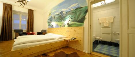 Een slaapkamer met een houten bed, een koperen vossenvel tapijt, een muurschildering van een berglandschap, een kleine tafel met een telefoon, en een badkamer met een geruit vloertje.