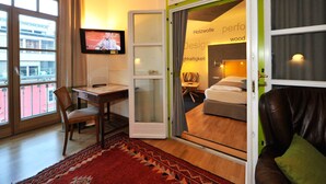 Habitación de hotel con cama, escritorio con silla, televisión y vista a la ciudad a través de amplias ventanas.