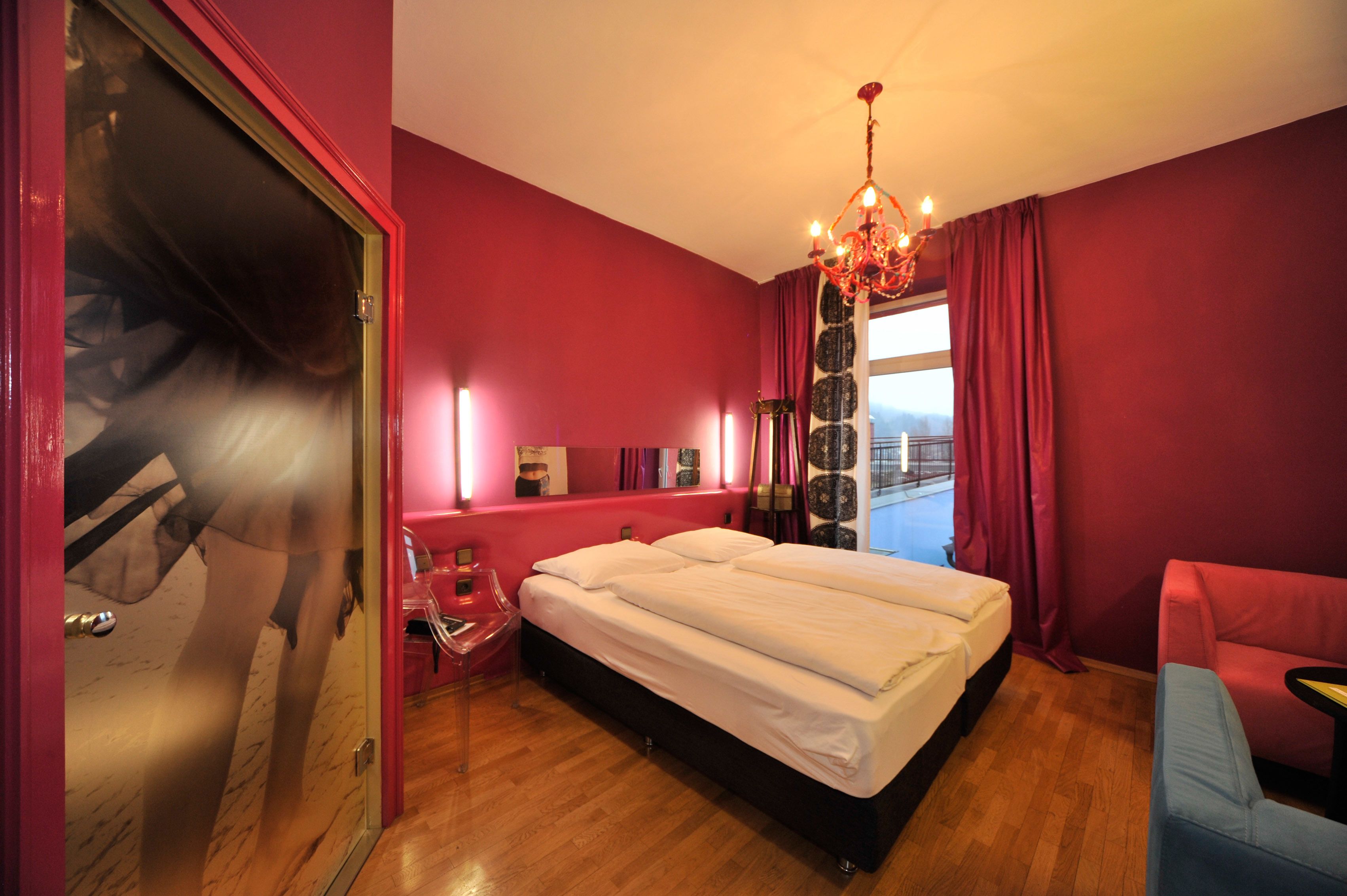 Ein Hotelzimmer mit einem großen Bett, einer roten Wand, einer Chandelier und Blick auf die Stadt.