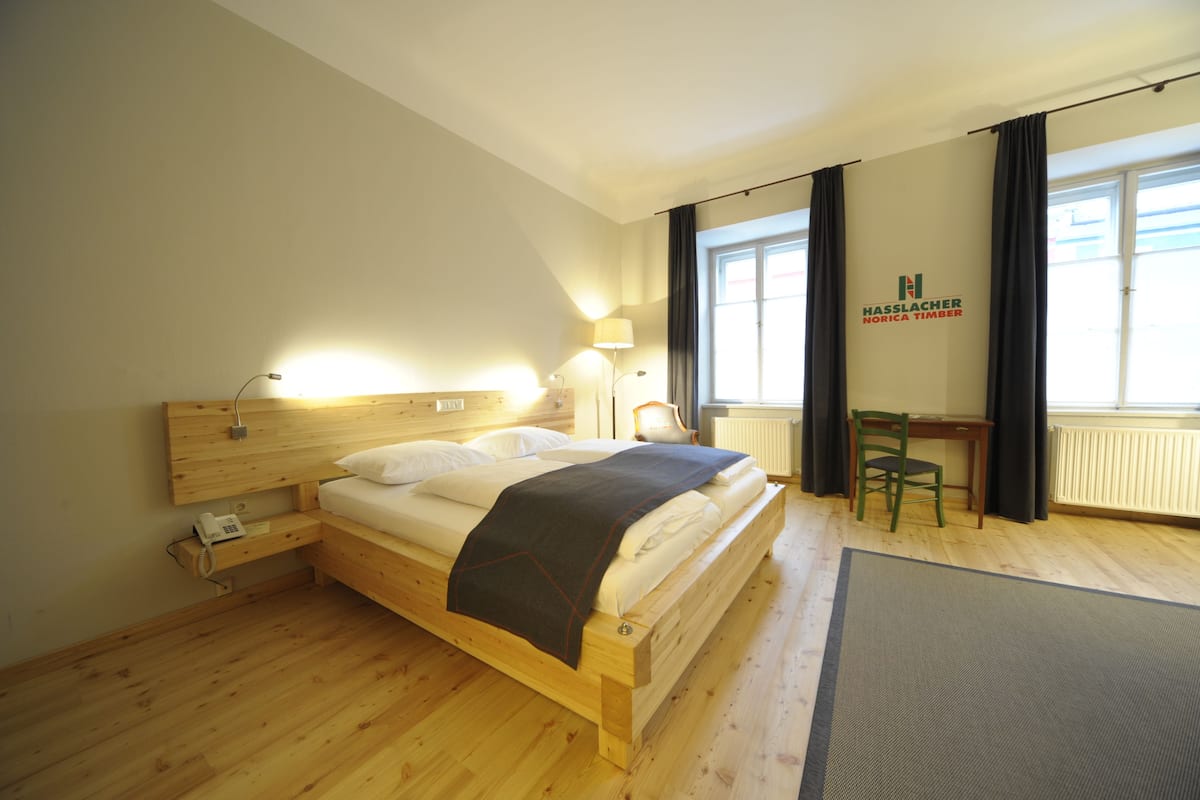 A wooden bed with a headboard, bedside lamps, a desk, and a chair in a room with large windows and a radiator.