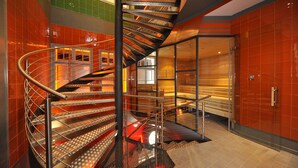 Una escalera espiral moderna con barandillas de metal y cerramiento de vidrio, rodeada de paredes de azulejos naranjas y un suelo pulido.