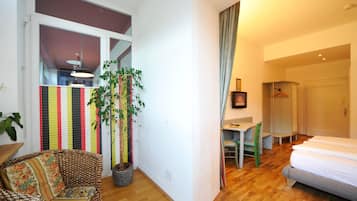 A compact hotel room with a bed, a small table, a chair, a potted plant, and a colorful striped partition.