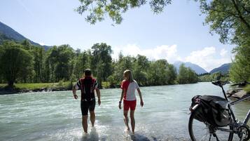 Zwei Personen laufen durch einen Fluss, in der Nähe steht ein Fahrrad.