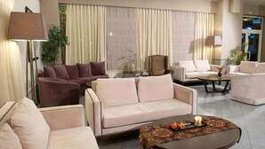 A modern living room with a beige sofa, a brown armchair, and a coffee table with a tray of fruit and a candle. - Aliakmon Hotel (Kozani)