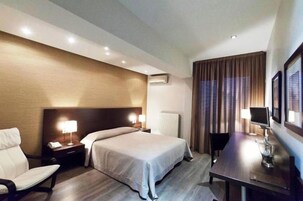 Una habitación de hotel con cama, mesitas de noche, una silla, un escritorio con lámpara y una ventana con cortinas.