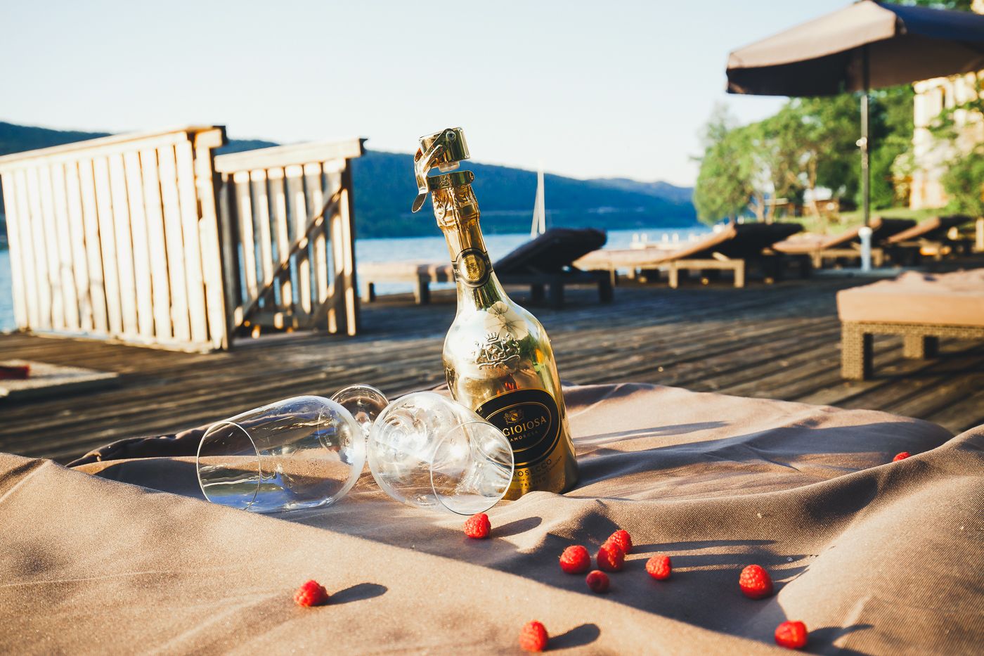 A bottle of wine, empty wine glasses, and scattered strawberries on a beach with wooden deck and lounge chairs.