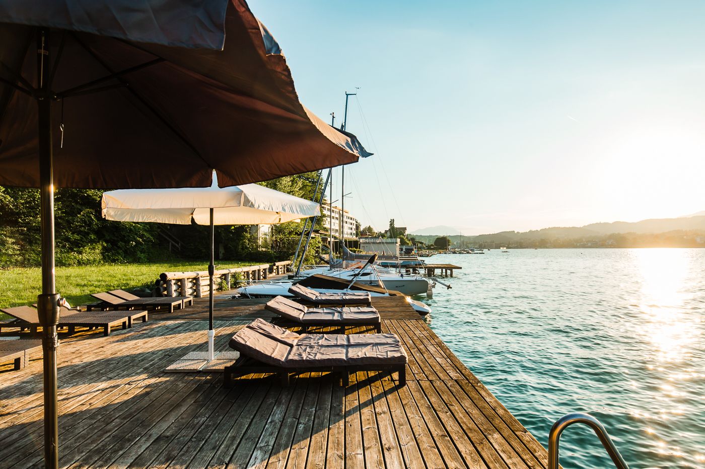 Une jetée en bois, agrémentée de chaises longues et de parasols, qui offre une vue sur une étendue d’eau calme, avec des bateaux et des bâtiments en arrière-plan.