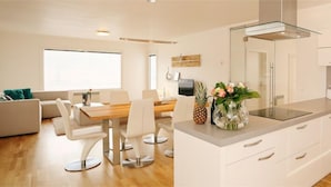 A modern kitchen with white cabinets, a stainless steel sink, and a built-in microwave. There is a dining area with a wooden table and white chairs.