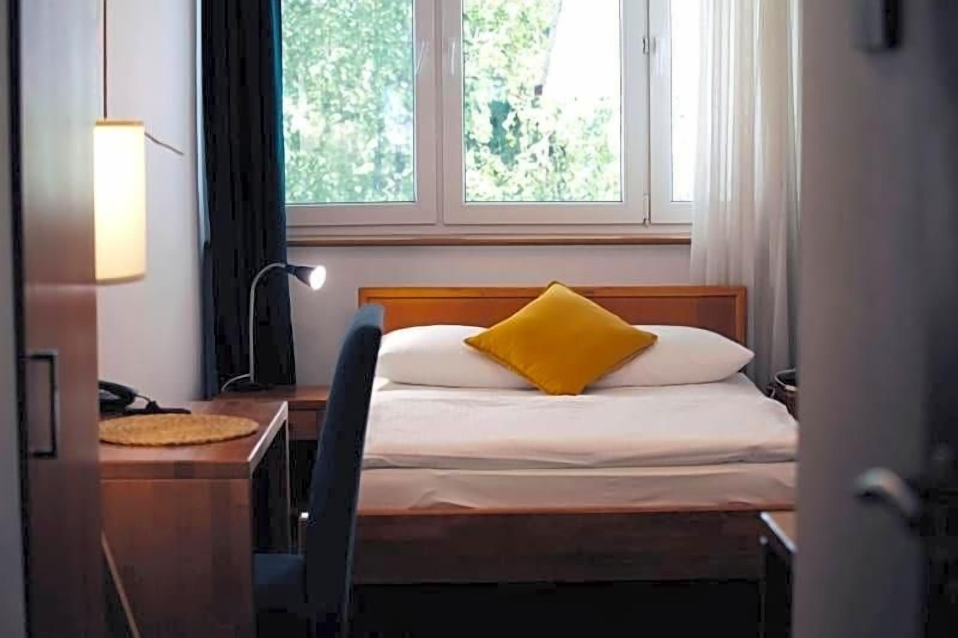A hotel room with a bed, a desk, a lamp, and a window with a view of trees.