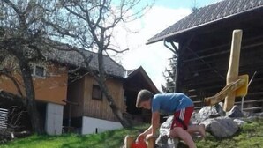 Une personne travaille dans un jardin, un tuyau d’arrosage à la main, entourée de gros rochers et, au fond, on aperçoit une structure en bois.