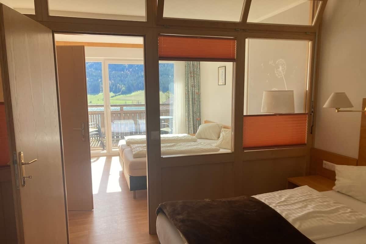 Una habitación de hotel con una cama grande, un balcón con vistas a zonas verdes y una zona de estar separada con una mesa y sillas.