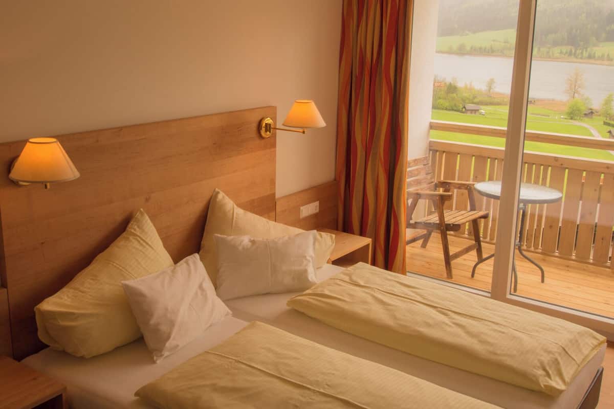 Habitación de hotel con cabecera de madera, dos camas con ropa de cama blanca, un balcón con mesa y sillas, y vistas a un lago y vegetación.