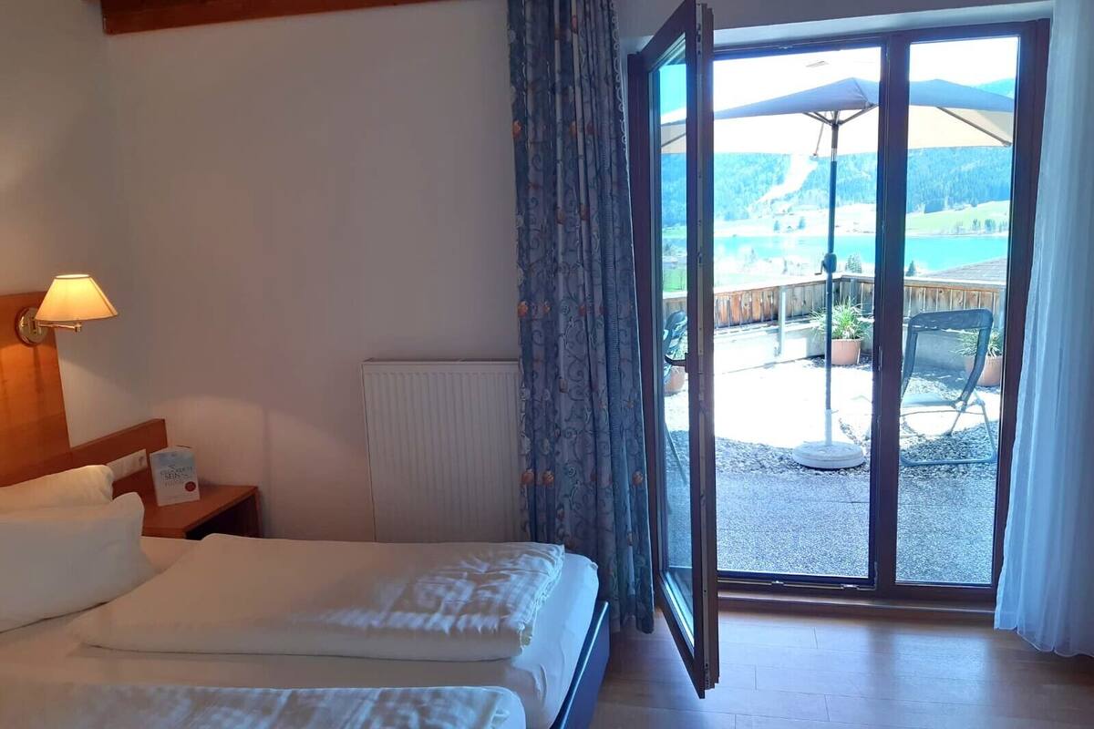 Habitación de hotel con cama, mesita de noche, lámpara y vista a un patio con mobiliario exterior.