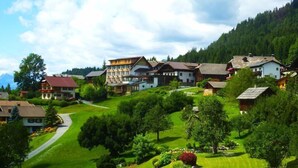 Un village avec des maisons construites sur une colline, entouré de verdure et avec des montagnes en arrière-plan.