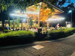 Un espacio de comedor al aire libre con mesas y sillas, rodeado de follaje y cubierto por una estructura con luces colgantes.