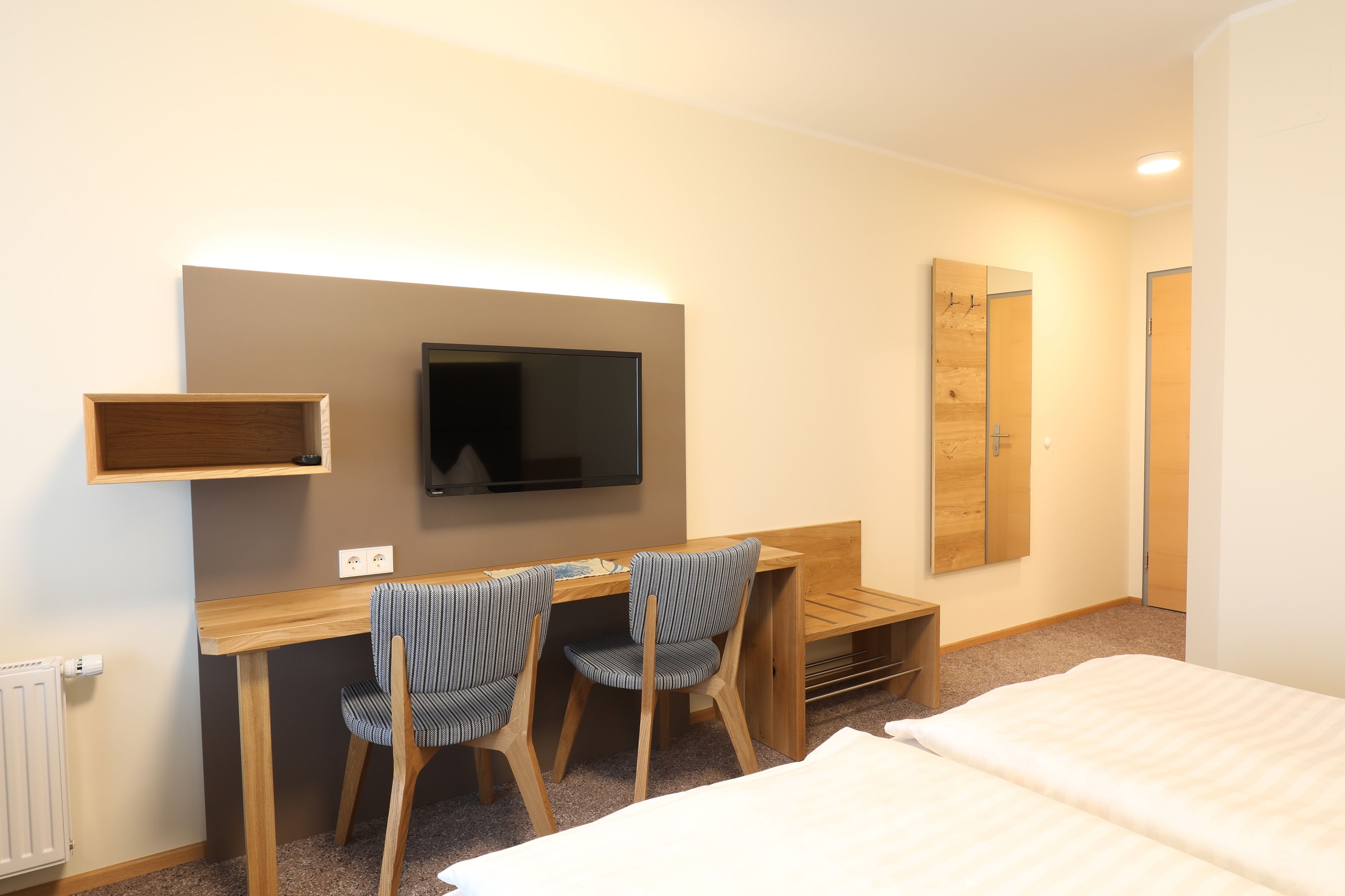 A hotel room with a wooden desk, two striped chairs, a flat-screen TV, a mirror, and a bed.