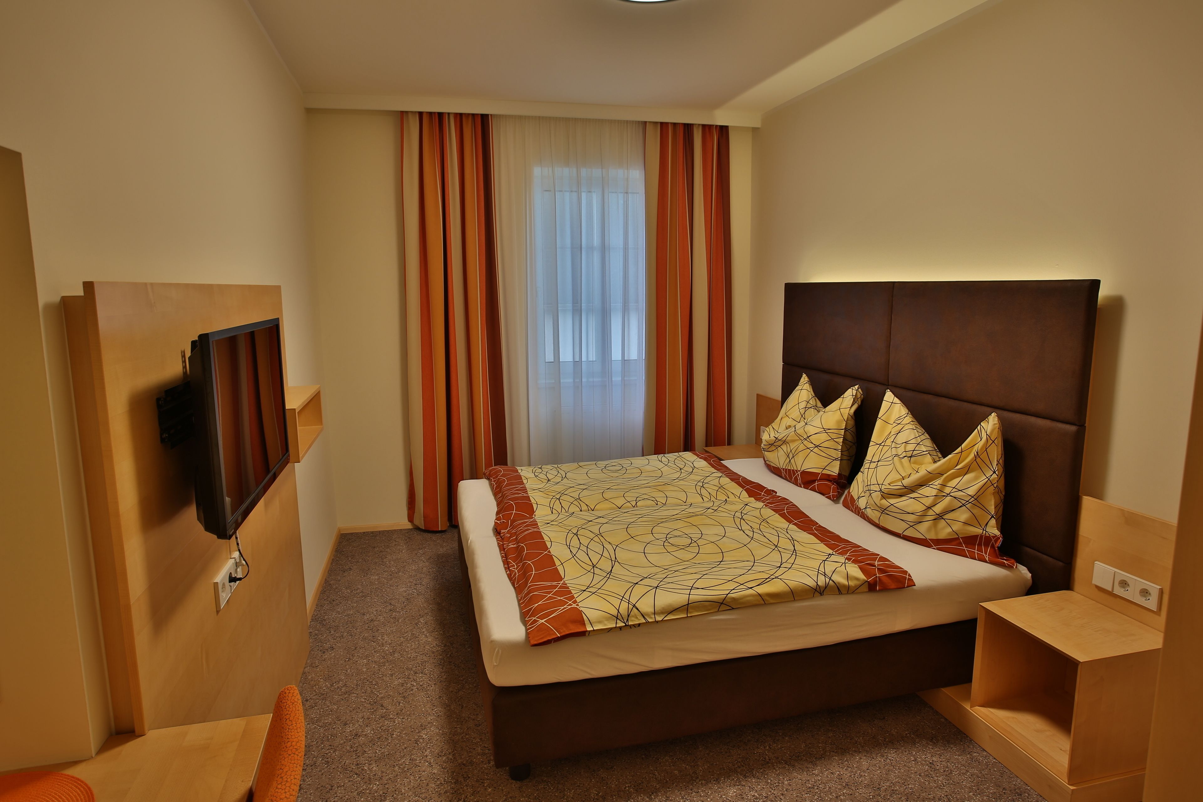 Habitación de hotel con una cama, televisor montado en la pared y ventana con cortinas.