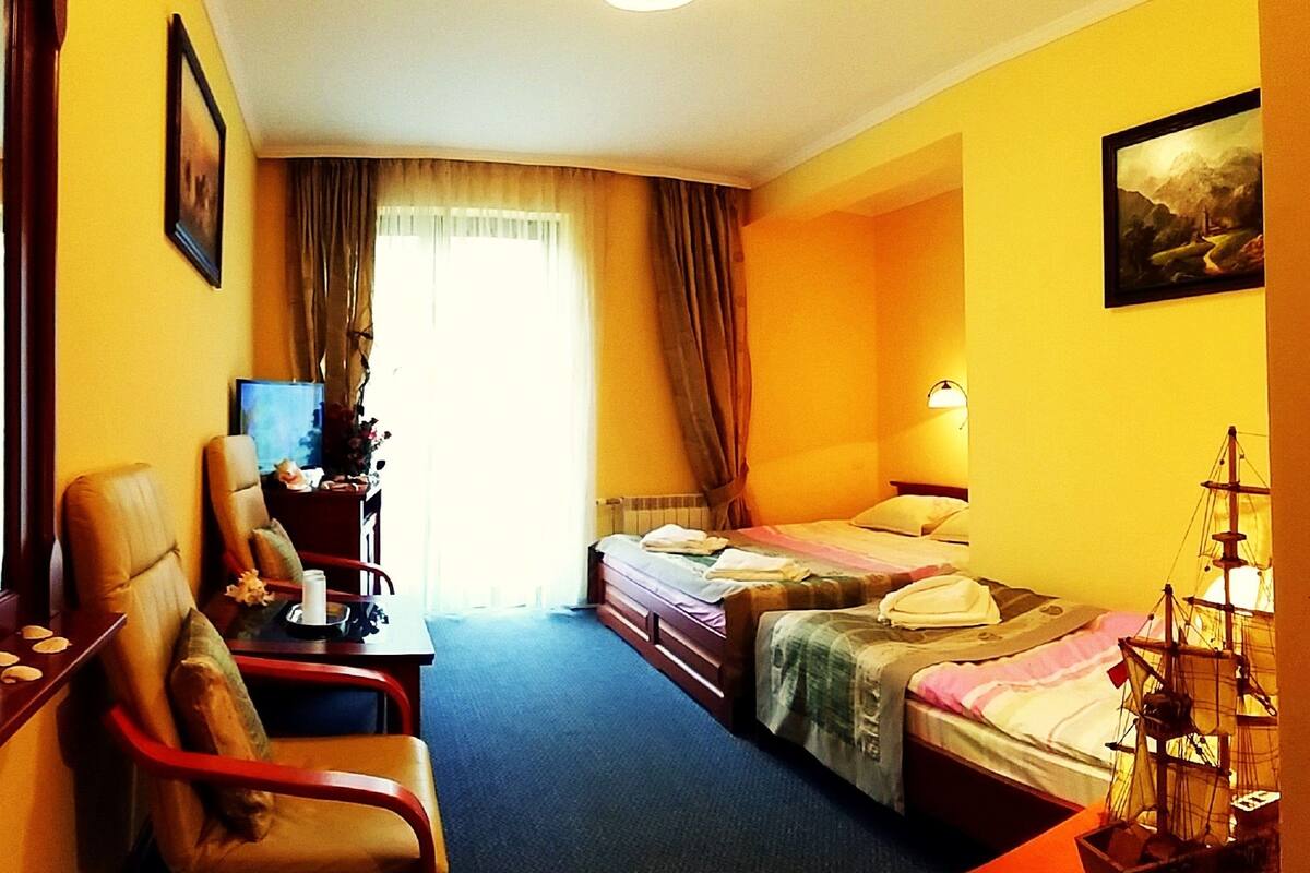 A hotel room with two beds, a desk with a computer, a chair, and a ship model on the nightstand.