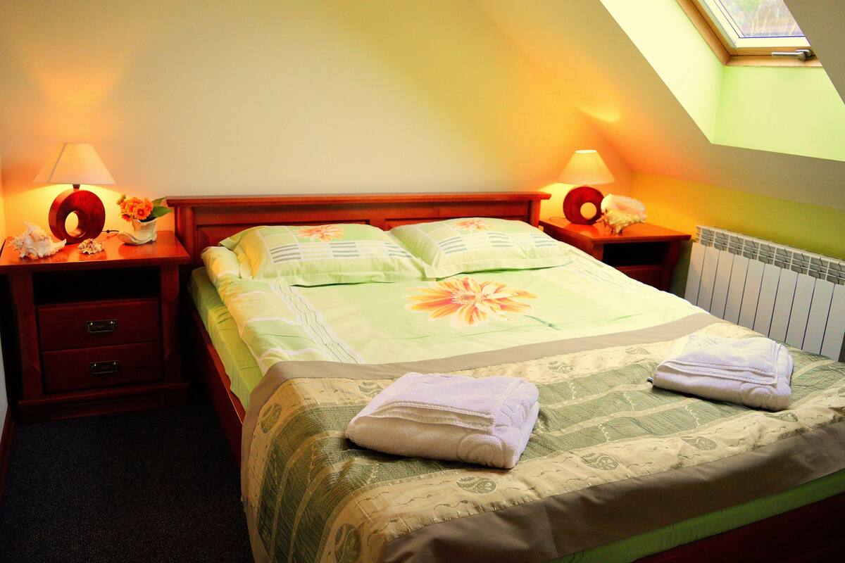 A neatly made bed with a floral bedspread, two pillows, and two folded towels on a wooden headboard. There are two bedside tables with lamps and decorative items.