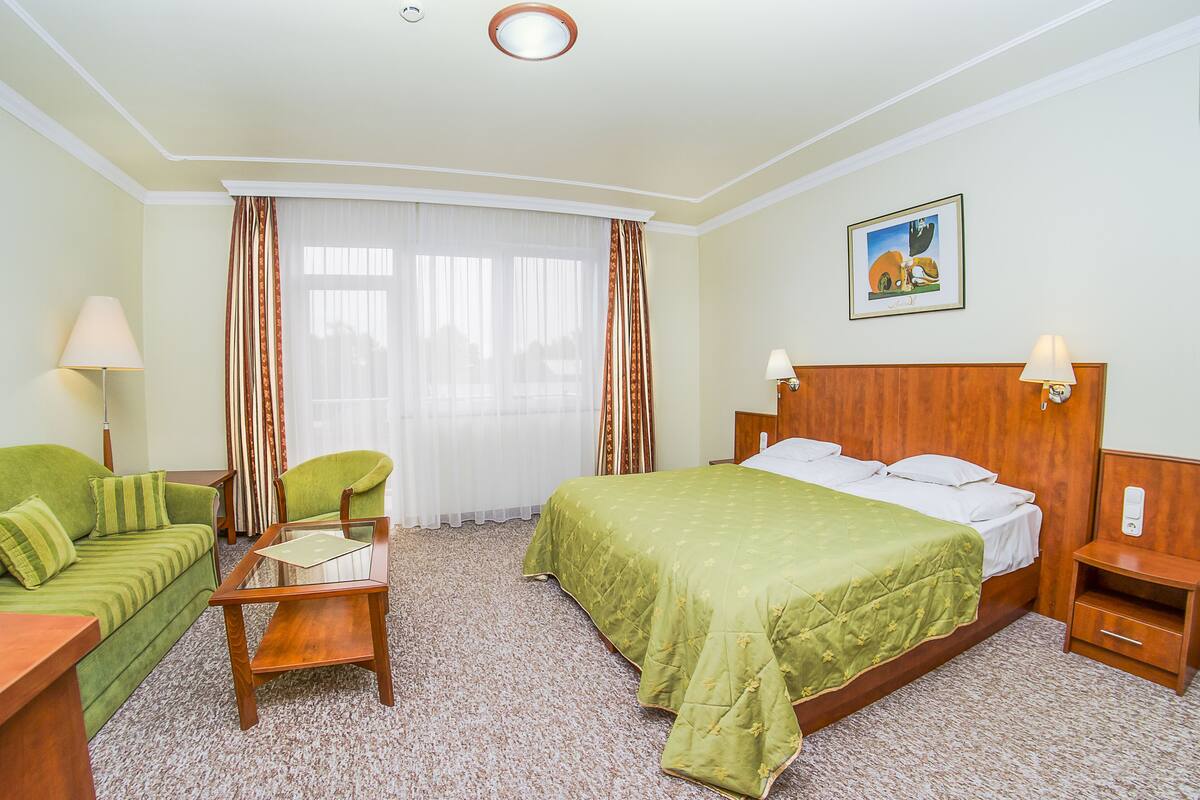 Una habitación de hotel con cama, sofá verde, mesita, mesita de noche y lámpara.