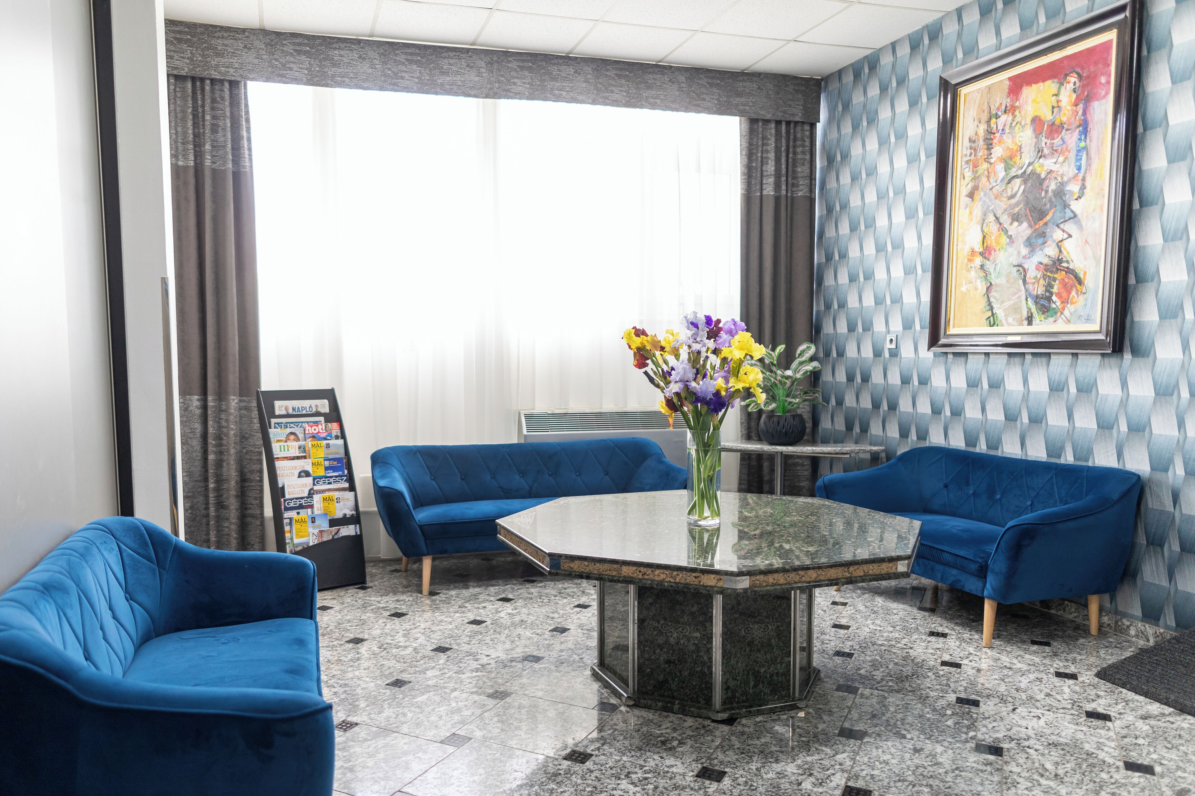 Un vestíbulo con dos sofás azules, una mesa de centro de vidrio, un jarrón con flores, un cuadro abstracto grande y un tablón de anuncios con varios objetos.