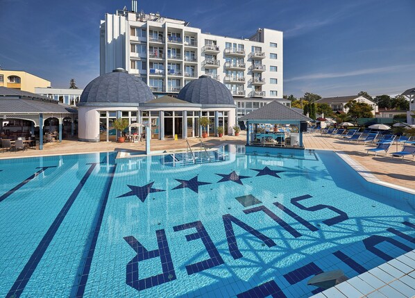 A swimming pool with star patterns and the word "SILVER" on the tiles, surrounded by lounge chairs and a building with multiple balconies. - Hotel Silver (Hajduszoboszlo)