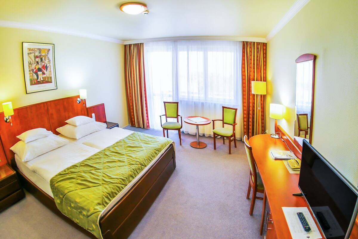 Habitación de hotel con una cama, dos sillas, un escritorio con computadora y una ventana con cortinas.