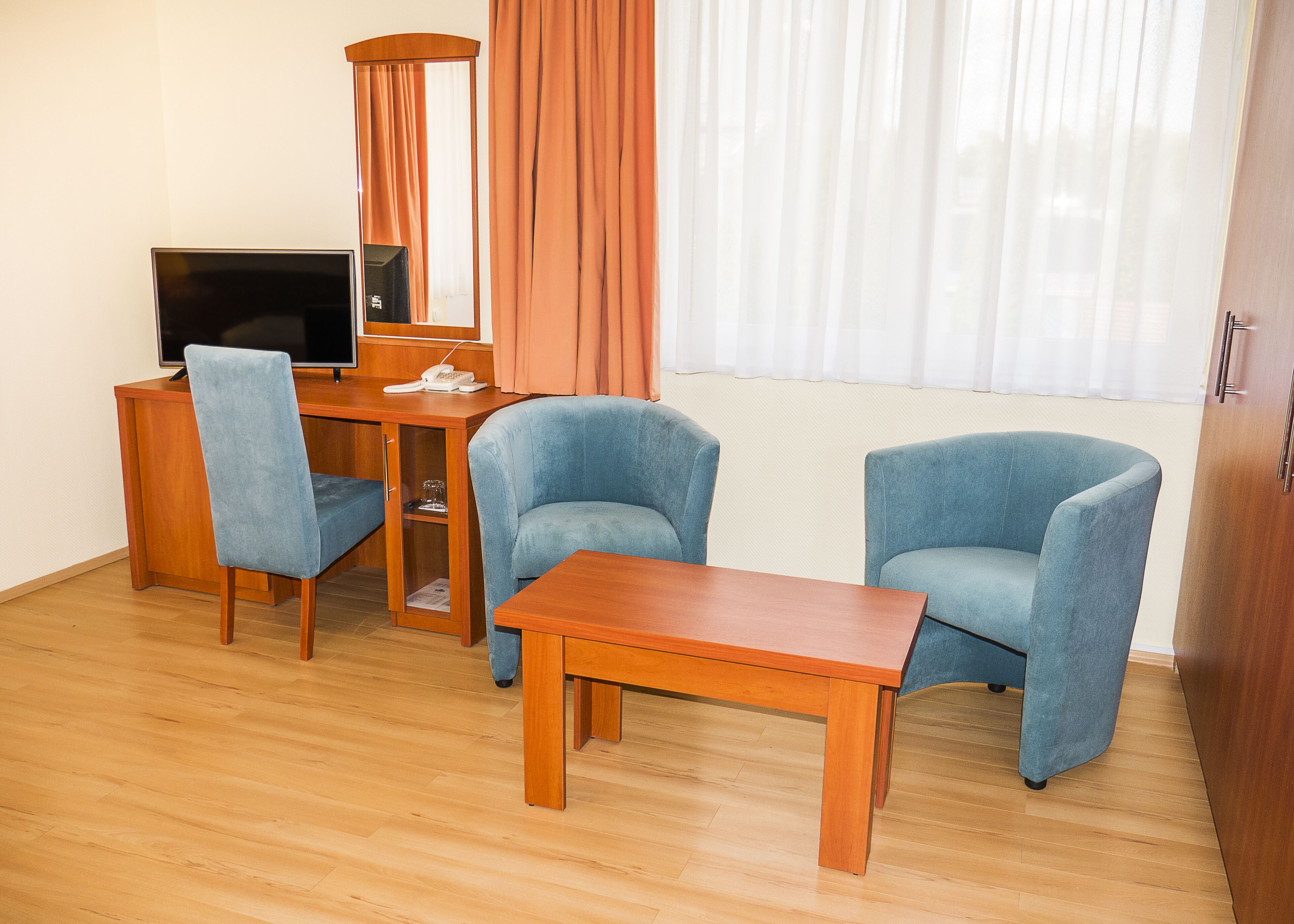 Habitación de hotel con un escritorio de madera, dos sillones azules, una televisión y una ventana con cortinas.