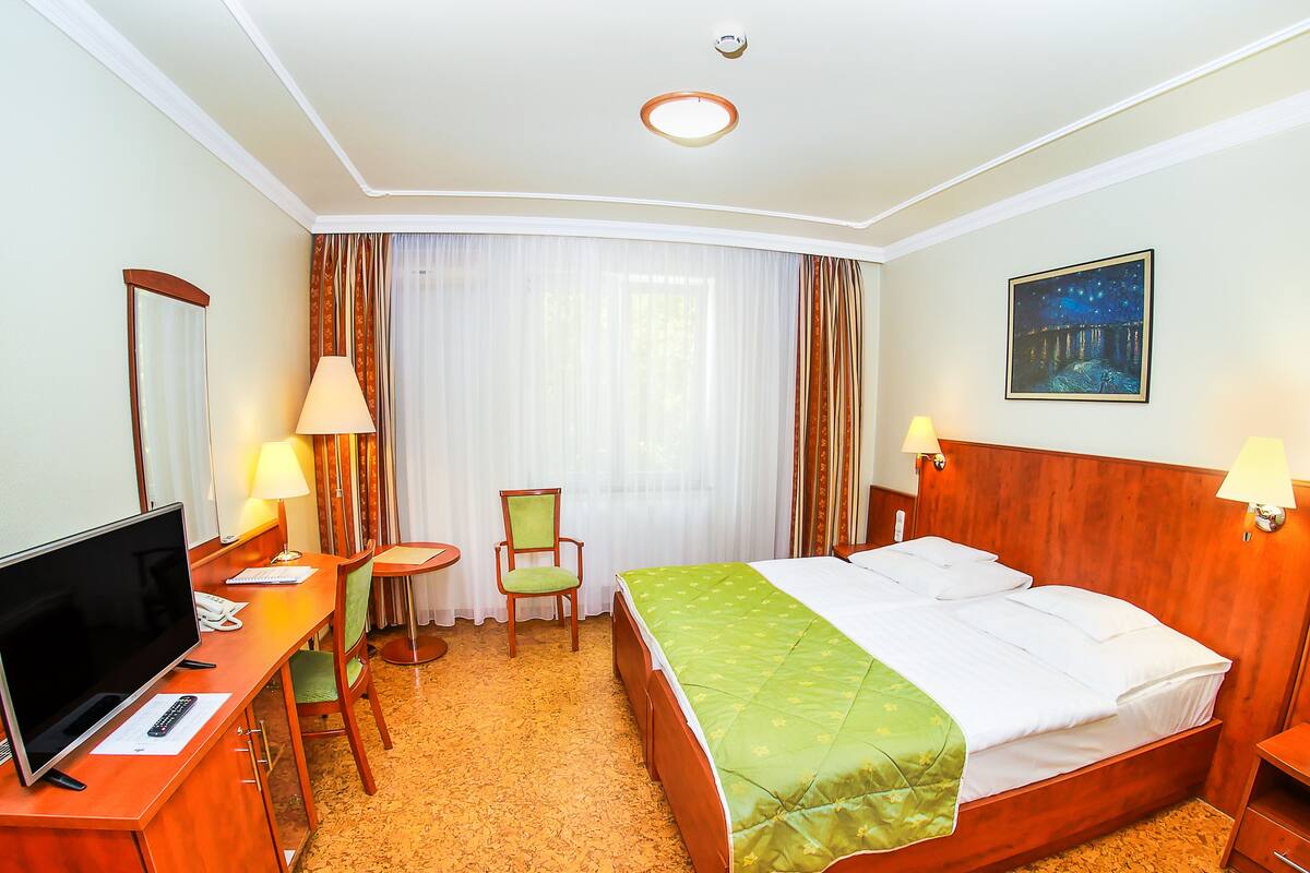 Habitación de hotel con cama, escritorio con televisor, una silla, una mesita y una ventana con cortinas.