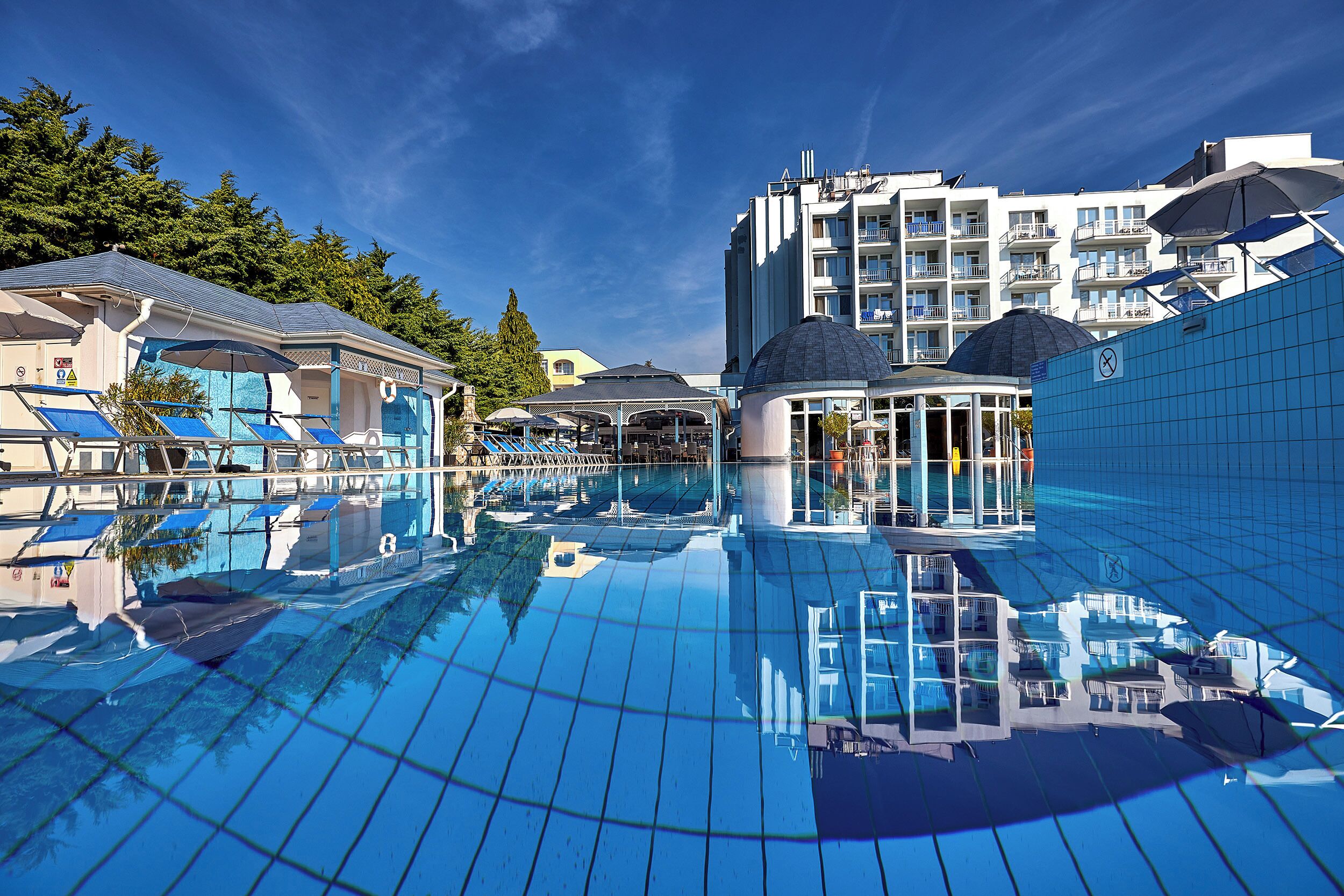 A swimming pool with clear blue water, surrounded by buildings with balconies and a covered area.