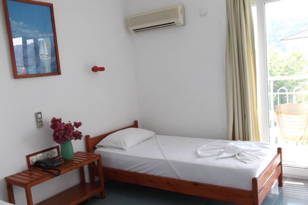 Una cama individual con estructura de madera, ropa de cama blanca, una mesita de noche con teléfono y jarrón, un equipo de aire acondicionado y una ventana con cortinas.