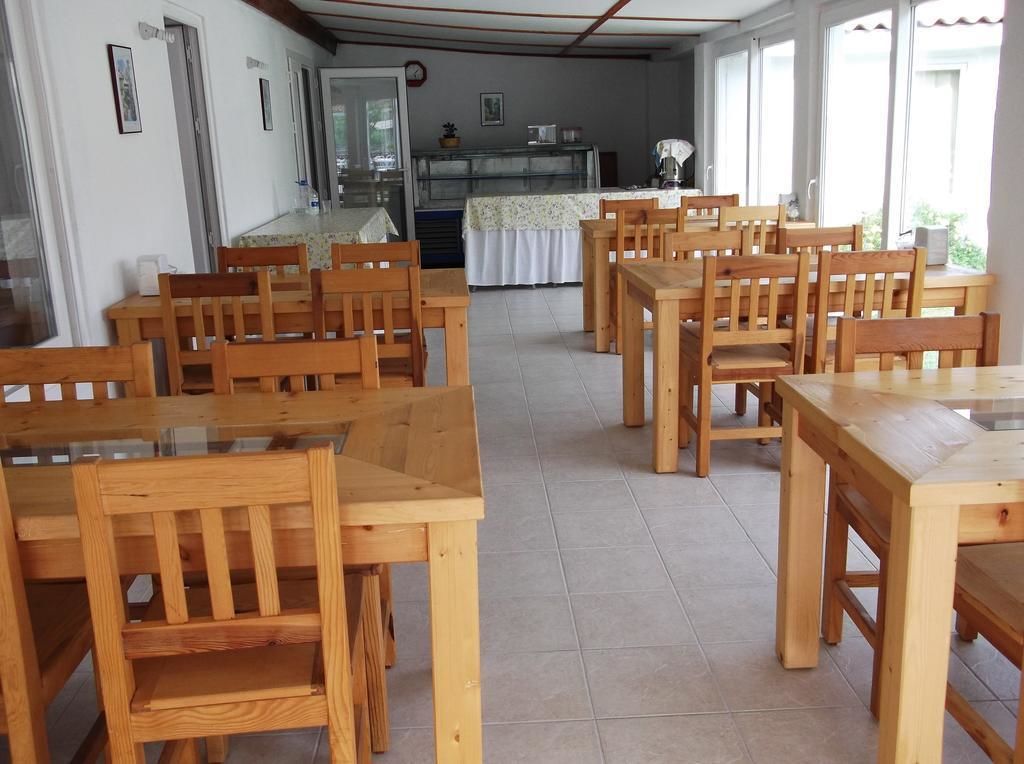 Uma sala de jantar com mesas e cadeiras de madeira, uma cozinha ao fundo, e grandes janelas que proporcionam luz natural.