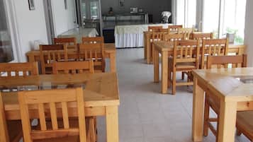 Uma sala de jantar com mesas e cadeiras de madeira, uma cozinha ao fundo, e grandes janelas que proporcionam luz natural.