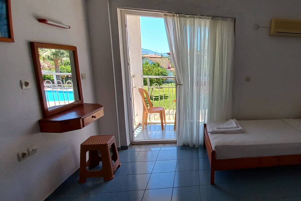 Un dormitorio con cama, un taburete pequeño, una mesita de noche, un ventanal con vista a un balcón y un equipo de aire acondicionado.